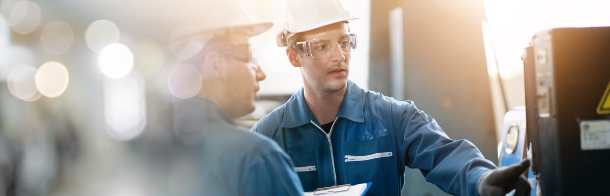 Two industrial workers in hard hats review machinery, one pointing at a control panel while the other holds a clipboard.