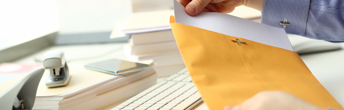 A person who is sitting at a desk is opening up a large envelope and pulling a piece of paper out of it.