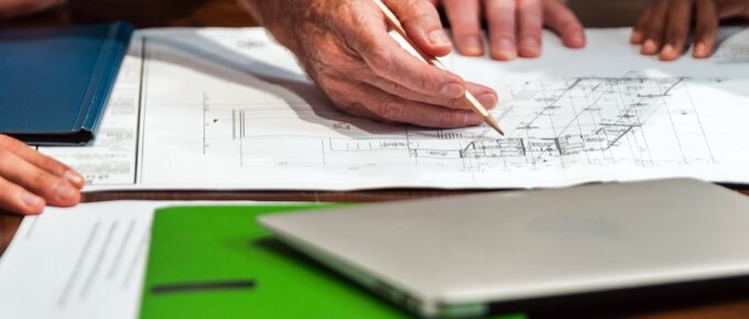 Several professionals are standing at a table, reviewing engineering schematics with a laptop and a clipboard nearby.