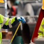 Two people, both of whom are wearing high-visibility jackets, are standing next to industrial equipment.