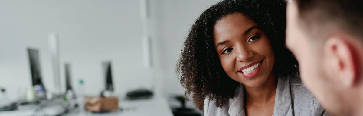 An over-the-shoulder view shows a woman wearing a gray blazer sitting at a desk, holding a pen and talking to a man.