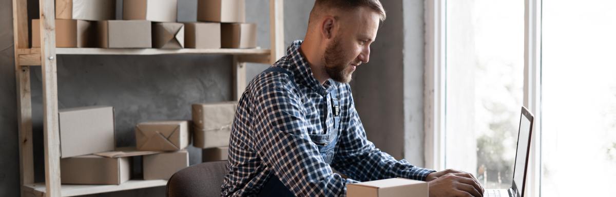 A businessman sits at his desk at home with packaging on the shelves behind him. He's working on the laptop.
