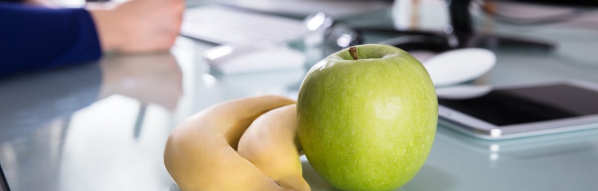 A couple of yellow bananas and a green apple resting on a desk beside someone working at their computer.