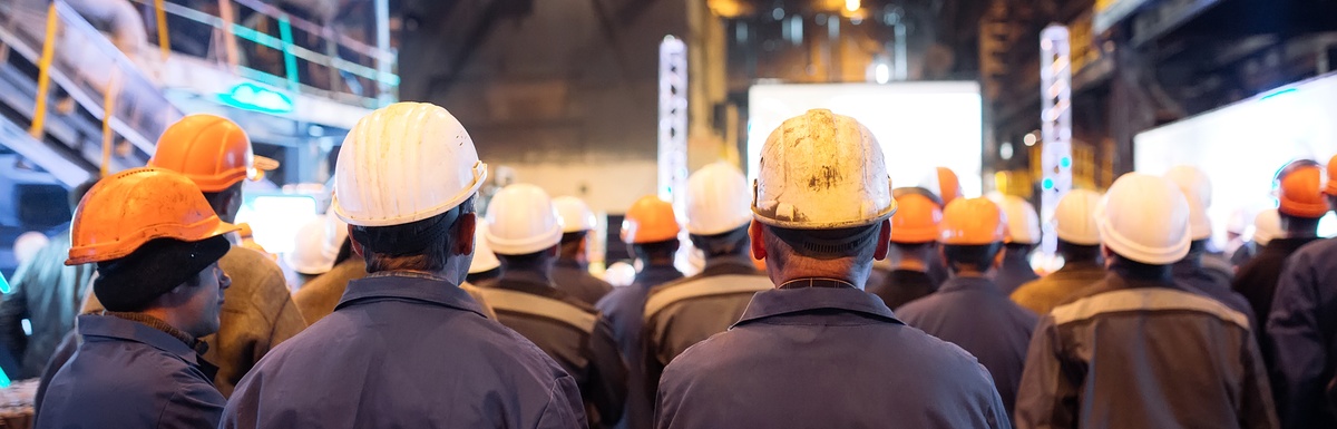 A group of workers in hard hats standing with their backs to the camera inside a large industrial facility.