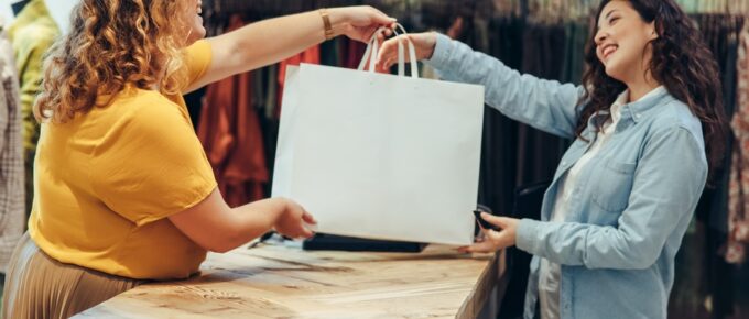 Two women stand opposite each other in a retail setting. The woman behind the counter hands a white bag to the other woman.