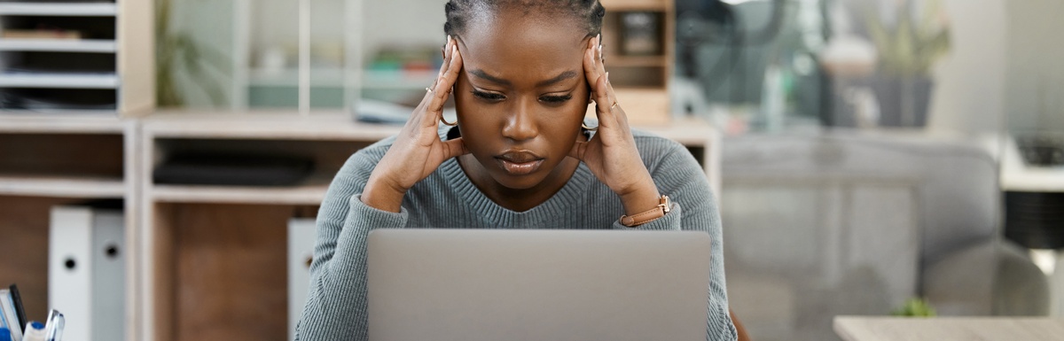 A professional sitting at their work desk and holding either side of their head while staring down at a laptop.