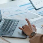 Two business professionals sit at a desk with a calculator, laptop, and a tablet to review a pie graph and data.
