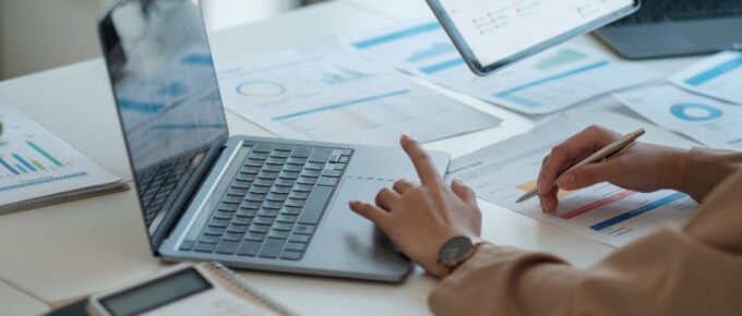 Two business professionals sit at a desk with a calculator, laptop, and a tablet to review a pie graph and data.