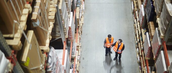 Two workers wearing safety vests standing in a warehouse aisle between tall shelves stacked with boxes.