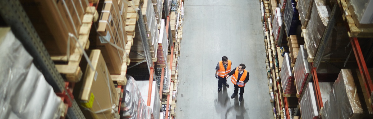 Two workers wearing safety vests standing in a warehouse aisle between tall shelves stacked with boxes.