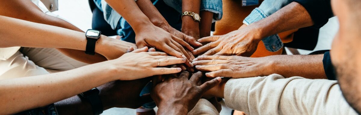 A group of people stacking their hands together while wearing casual clothing in a bright indoor space.