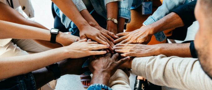 A group of people stacking their hands together while wearing casual clothing in a bright indoor space.