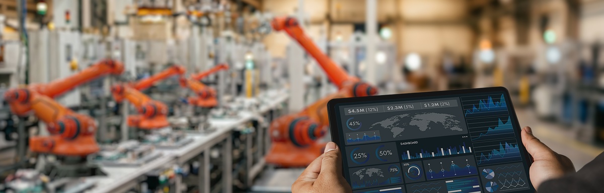 A person is holding a tablet in their hands as they stand in a warehouse. There is industrial equipment in the background.