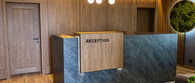 A lobby with a reception desk. The desk is mostly made out of wood and there are lights above the desk.