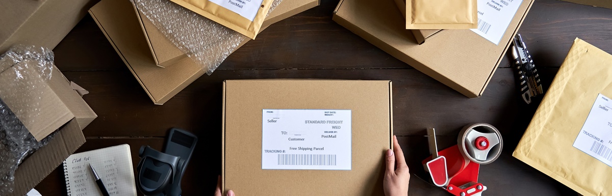 a pair of hands holding a labeled cardboard box on a dark table with mailers, bubble wrap, and a tape dispenser.