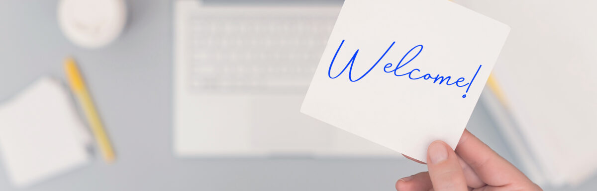 A new employee stands over their company laptop as they hold a "welcome" note over their workstation.
