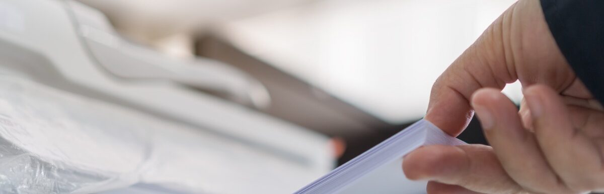 A close-up of a person lifting a stack of white paper from a printer tray in a bright office, with a blurry background.