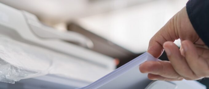 A close-up of a person lifting a stack of white paper from a printer tray in a bright office, with a blurry background.