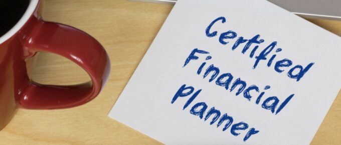 A close-up of a white sticky note with "Certified Financial Planner" written on it next to a pen and coffee mug.