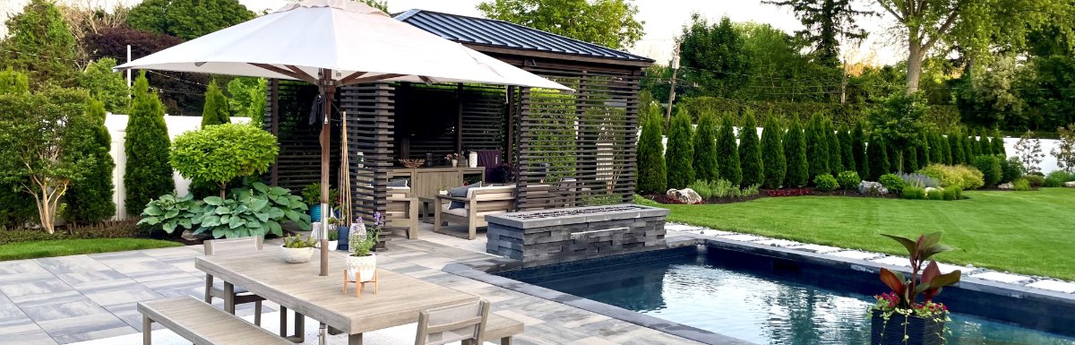A landscaped backyard with a swimming pool, wooden dining table, white umbrella, and covered seating area on a patio.